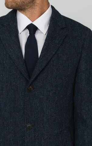 Close-up of a blue herringbone tweed overcoat worn with a white shirt and navy knitted tie.