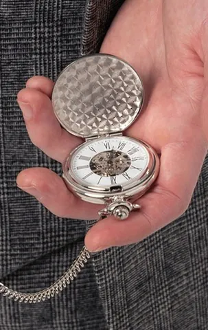 Open silver-coloured skeleton pocket watch with Roman numerals, held against a checked suit.